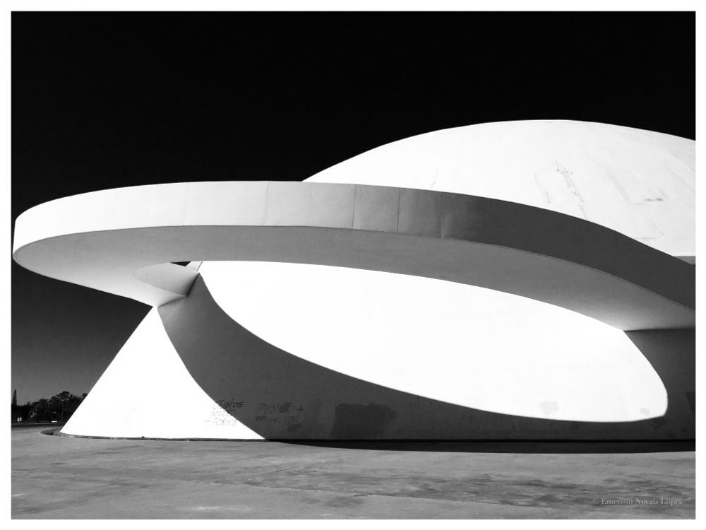 Photography. Black and white. The striking geometric lines of Oscar Niemeyer’s National Museum of the Republic, in Brasília. The white dome and curved external arched bridge promenade contrast with the dark cloudless sky.