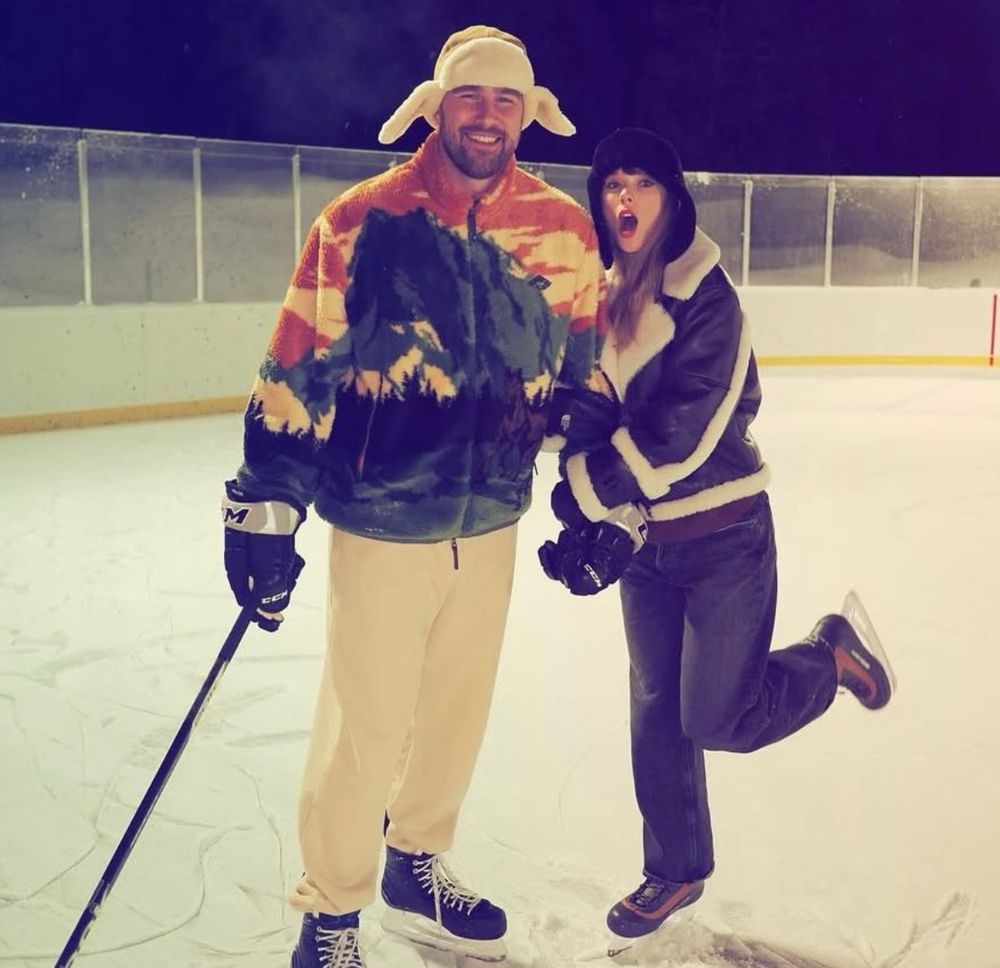 Taylor Swift and Travis Kelce both wearing ice skates in the middle of an ice rink. Travis is holding a hockey stick