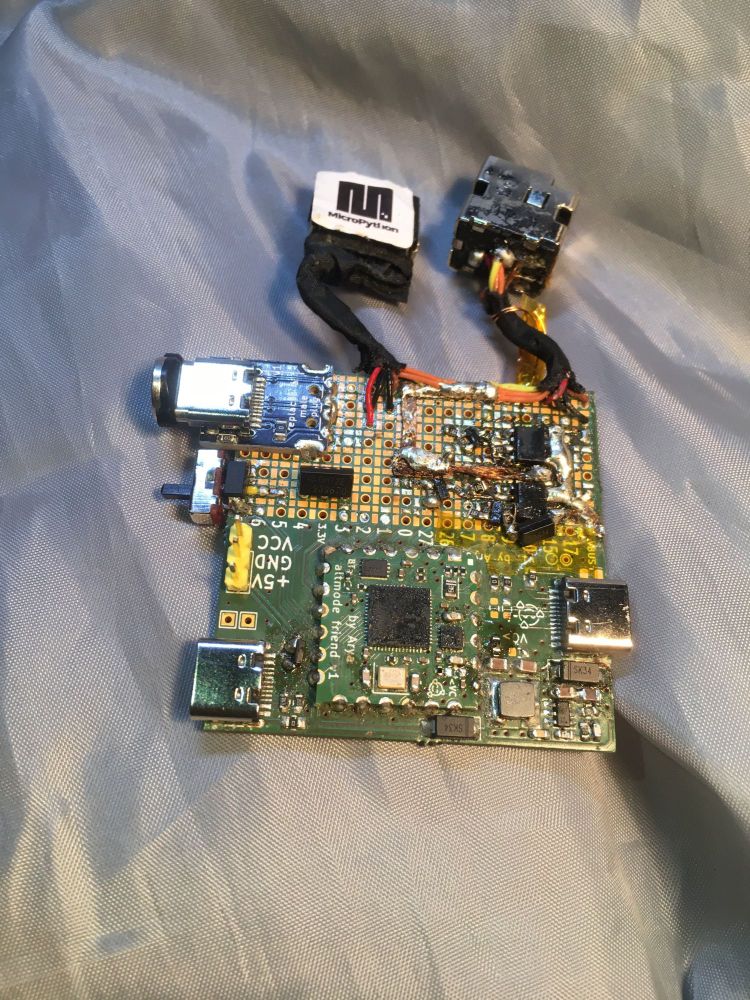 Picture of a USB-C RP2040-based devboard, with a prototyping area that a large amount of SMD parts is soldered onto, and there's two laptop power sockets attached to it. The devboard has also been modified in a few spots, some components substituted for others, traces cut and so on.