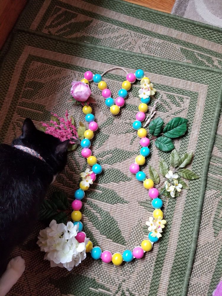 A wooden wall are piece of a silhouette of a bunny. It's painted yellow, pink, and blue, with fake flowers glued onto it.