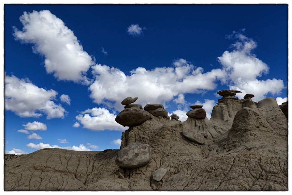 A group of oddly-shaped hoodoos emerges from the top of an eroded hillside. The blue sky above and behind is graced with puffy white clouds.