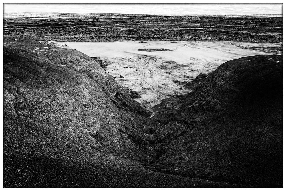 An image with sharp contrasts between the whitish upper-central triangle of a flat plain, and the much darker sloping humps of mudstone that surround it in the lower part of the picture. The horizon line is washed out/overexposed, as is the sky.