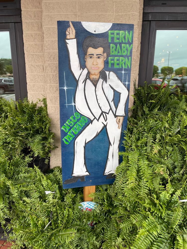 A large, unrealistic painting of John Travolta in his white disco suit from Saturday Night Fever is leaning against a wall, and is surrounded by potted ferns for sale. The sign also has text reading “FERN BABY FERN,” and “DISCO INFERNO.” Get it? Haha. 