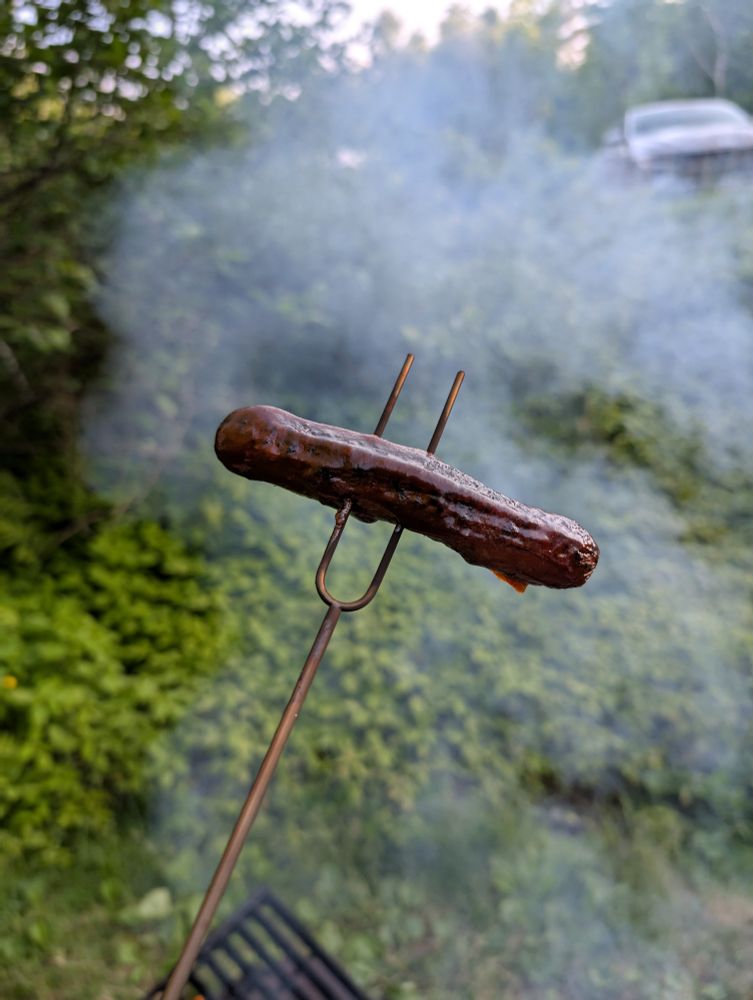 a roasted hot dog covered in barbecue sauce on an extendable camp fork. smoke from the campfire is visible in the background 