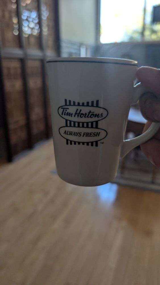 A white Tim Hortons coffee cup with the always fresh slogan hoisted in a blurred indoor mid century modern room. Grey natural light beans from a high clerestory window.