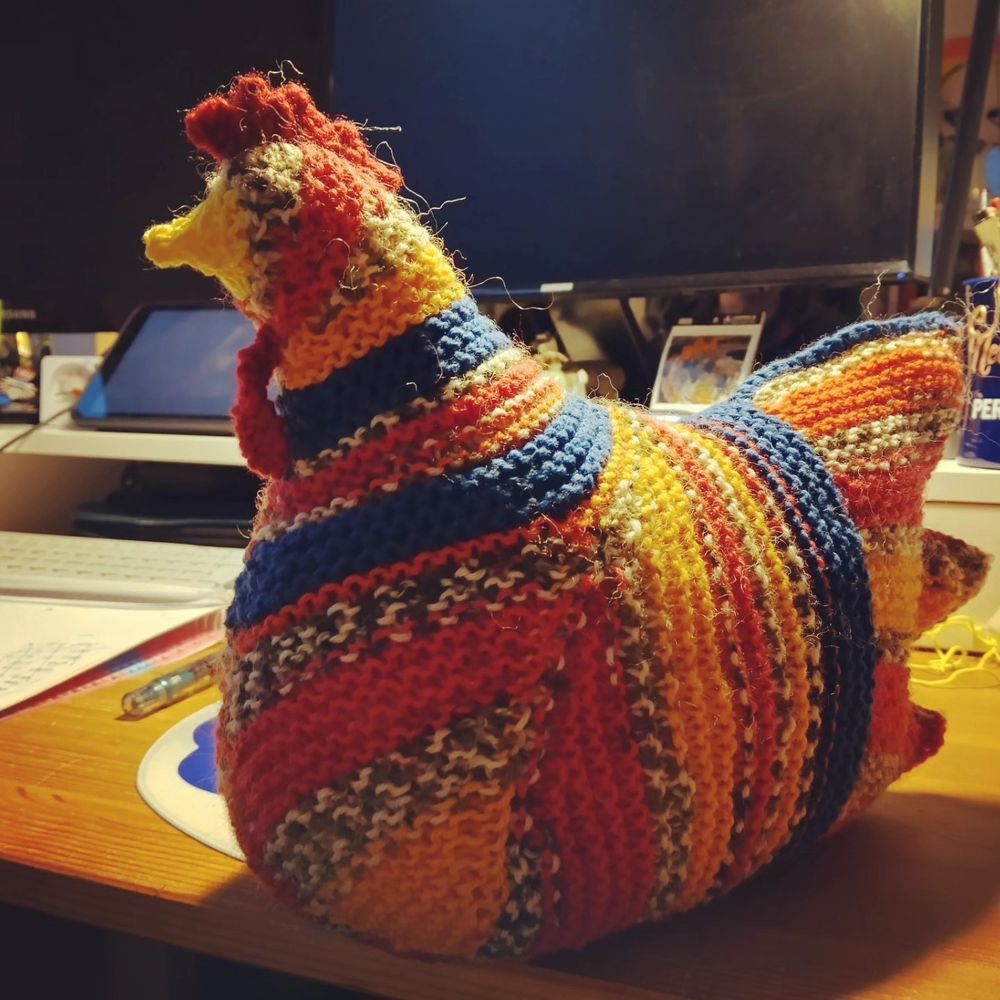 Knit chicken sitting on a desk. Chicken is red with melted grey and white stripes, orange stripes and blue strips. 