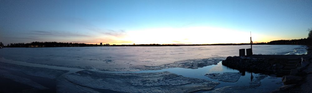 Panorama of a frozen bay at sunset, with a tiny patch of open water near a dock on the right-hand side.