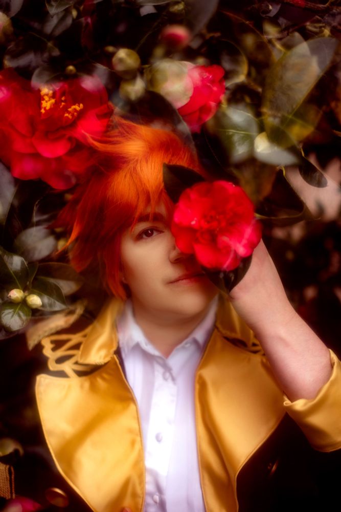 A photo of Marissa cosplaying Sylvain from Fire Emblem: Three Houses, standing in a red camellia bush and holding a bright red bloom that obscures one eye. Another photo of flowers from a different part of the bush are double exposed in the top left corner, creating an artsy magazine editorial vibe.