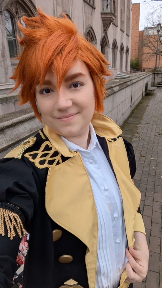 A selfie of Marissa in a Sylvain cosplay, a styled orange wig and the Garreg Mach academy outfit.