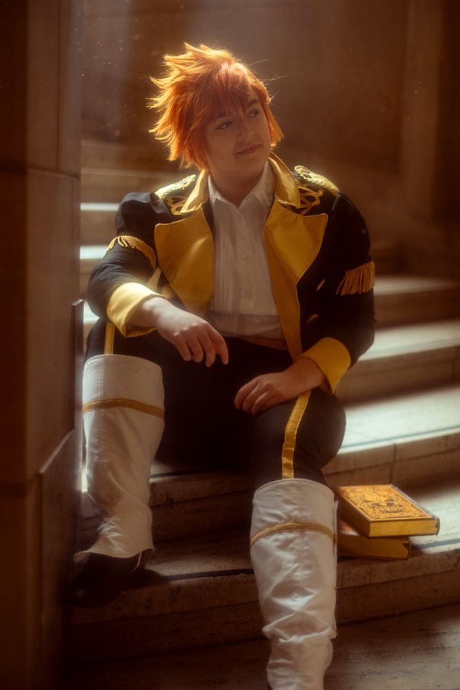 Photo of Marissa in Sylvain cosplay sitting on the steps of a spiral staircase, two books on a step below. She's looking to the right, sunlight reflecting off the back of her wig, highlighting the styling of Sylvain's hair.