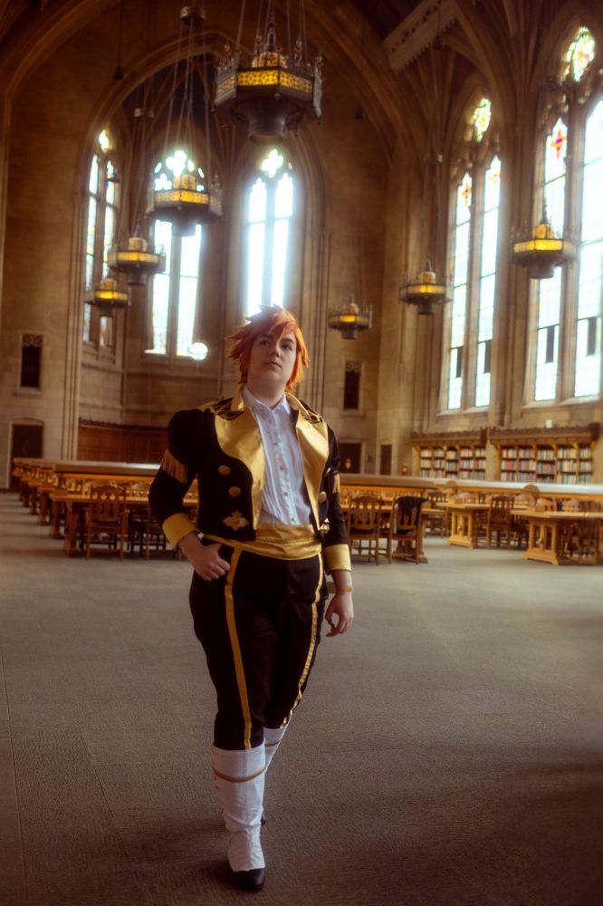 The same Sylvain cosplayer in the previous photo, strolling in the reading room. Large chandeliers hang overhead as light shines through large gothic windows.