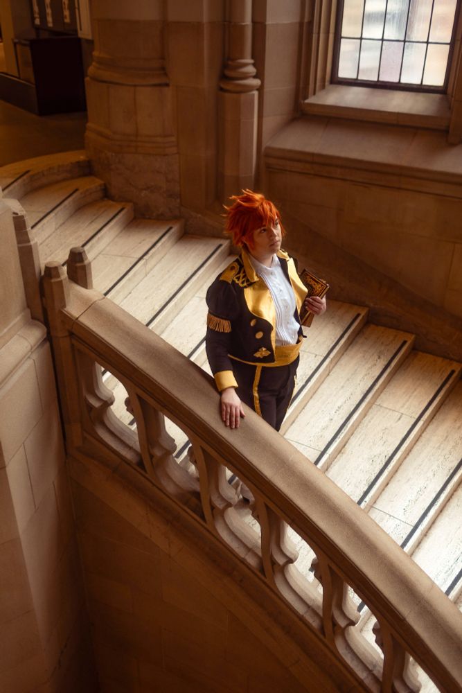 A high angle photo looking down at Marissa dressed as Sylvain from Fire Emblem: Three Houses walking down a spiral stairwell, book in hand.