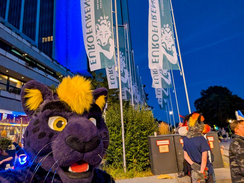 Finn, a purple and yellow panther fursuiter, posing in front of Eurofurence flag banners in the courtyard of the convention center