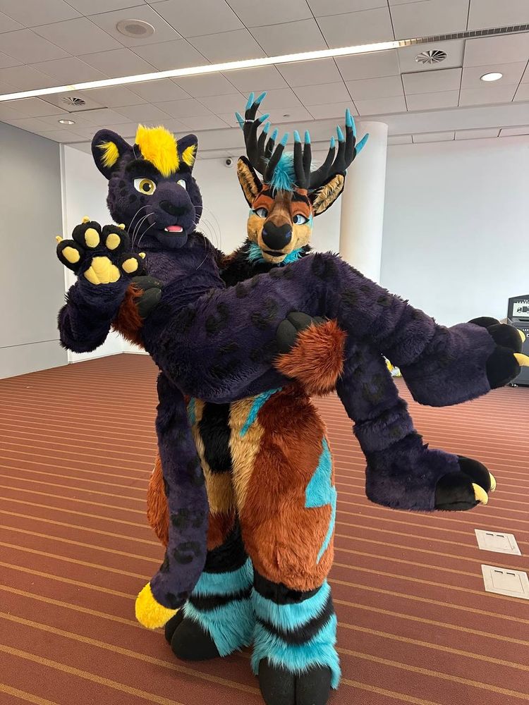 Volt, a brown and blue elk fursuiter, hoisting Finn, a purple and yellow panther fursuiter, in the air in the Anthrocon convention center