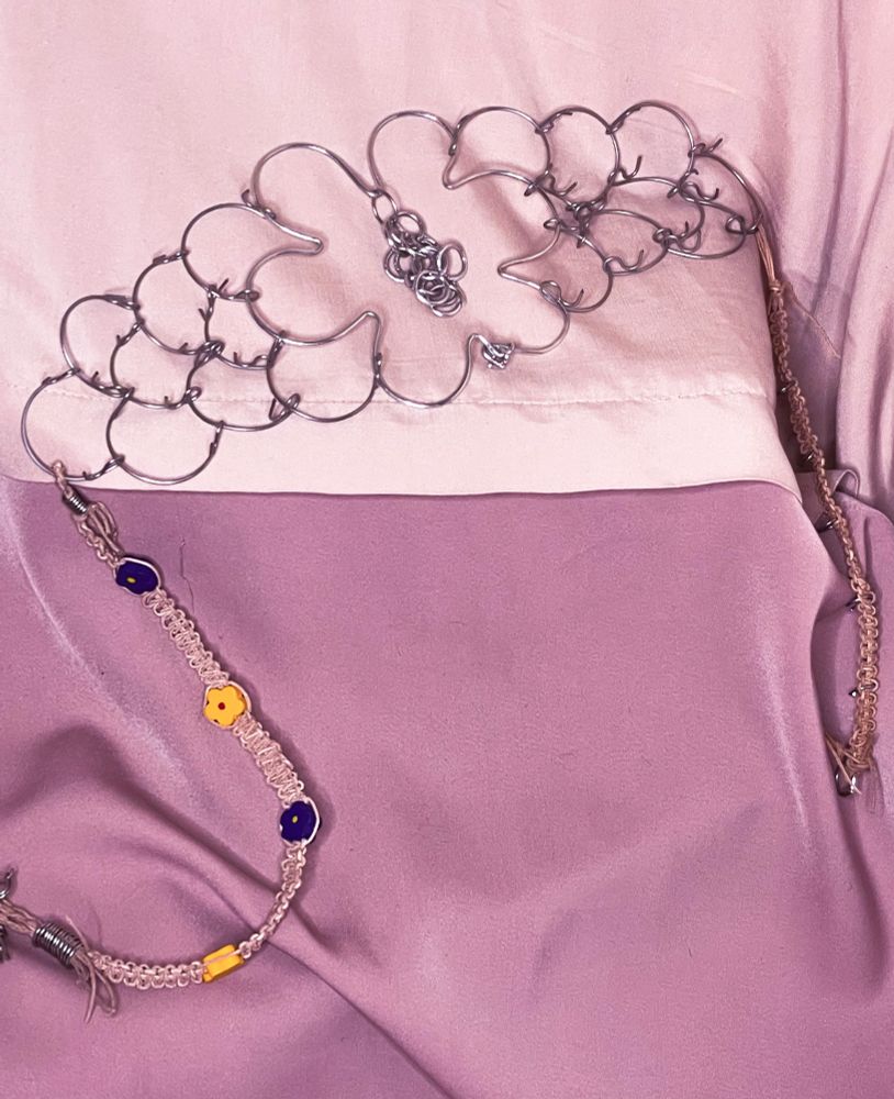 A three row stainless steel cilice with a flower shaped jump ring in the center, the flower has chains hang from the top center. The cilice has leather string weaved like friendship bracelets with purple and yellow wooden flower beads.