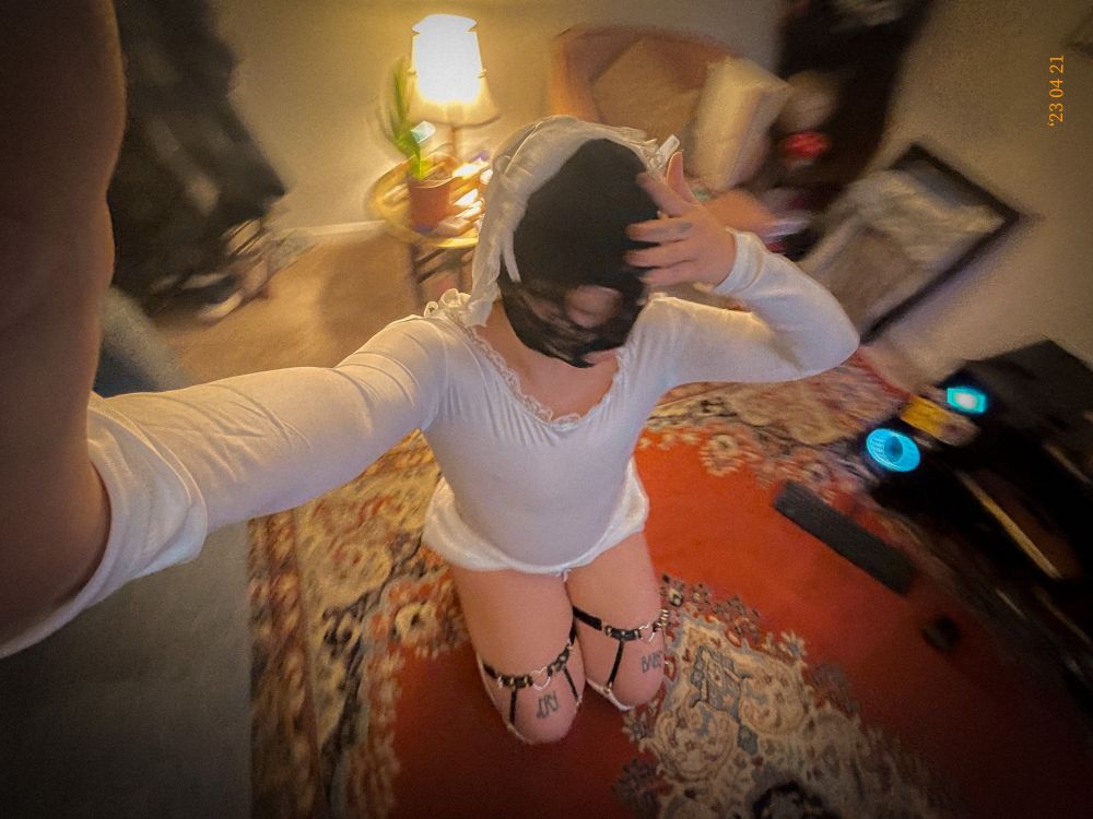 Noor kneels on the floor and looks away from the camera, a wide lens making their arm look long and the background slightly warped and there’s a blurr of motion. They wear a white body suit, fluffy white bloomers, black sock garters, and fluffy white socks. A black lace hood covers most of their face with holes for their eyes and mouth. 