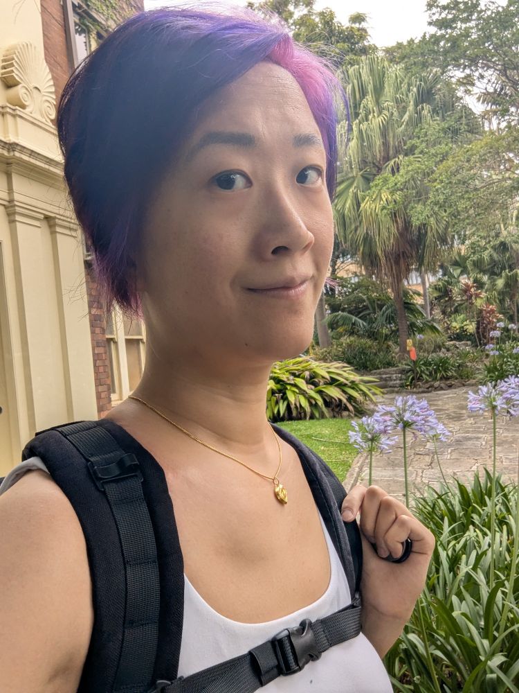 A girl with purple hair and a white sports bra wearing a rucking backpack. She's standing in front of a building on the left of the image and plants with purple flowers on the right.