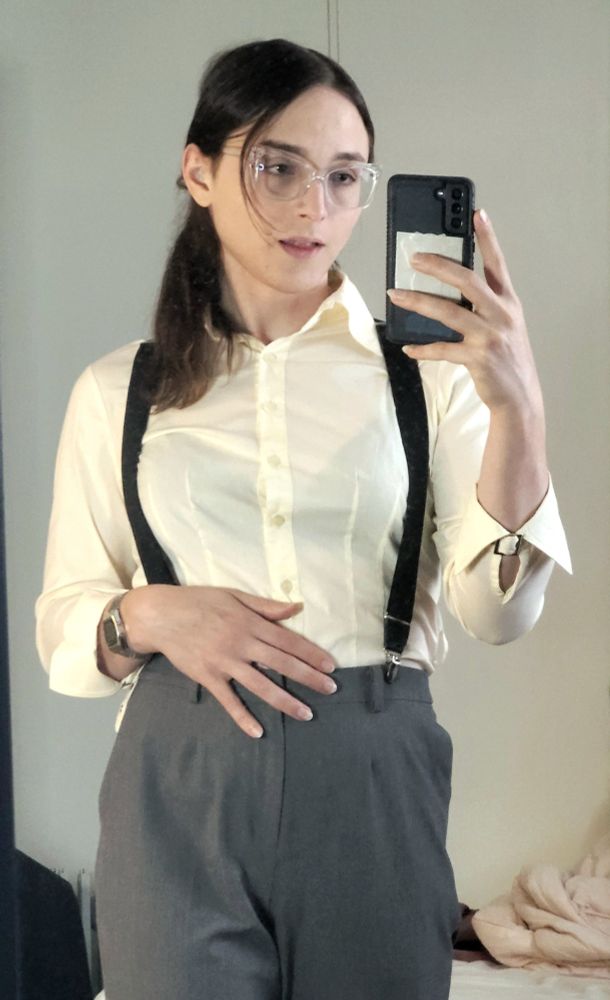 Mirror selfie of yoghurt with Grey dress pants held up by black suspenders over a tucked in cream colored button down with the sleeves partially rolled up,  casio marlin watch,  hair in ponytail and light makeup.  