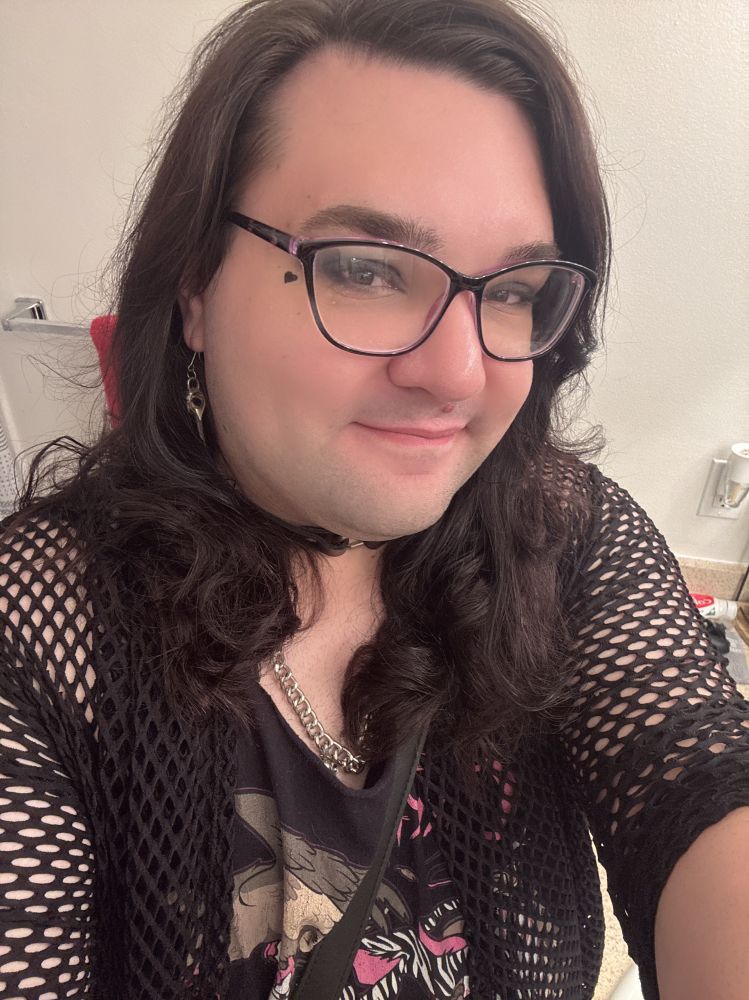 A selfie of an androgynous person smiling at the viewer with a fishnet top, black and varied colored tank top with silver jewelry and earrings, and wearing rectangular black/purple glasses.