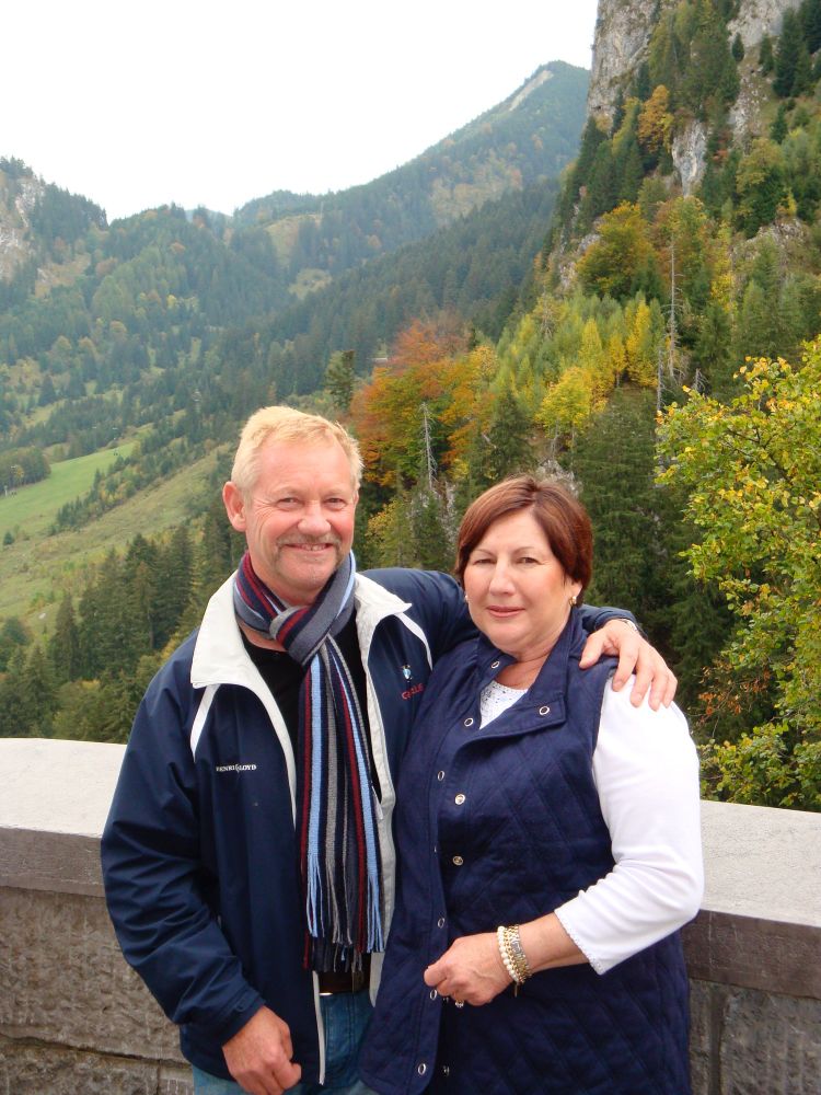 Two people stand on the ramparts of a castle. On the left is a blond man with a moustache, wearing a dark blue sailing jacket and scarf, smiling as he puts his arm around the woman next to him. She has reddish hair, and is wearing a sleeveless blue jacket and a white top. Behind them are verdant green forests. They look happy.