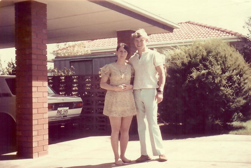 Two very young people stand just outside a brick carport, in front of a green bush. On the left is a woman in a short pale sundress; on the right, with his arm around her, is a taller young man wearing a white shirt and white slacks. They are both smiling happily.