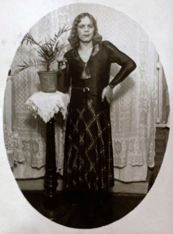 A black and white photograph shows a woman standing next to a pedestal holding a pot plant, in front of white lace curtains. The woman has shoulder-length curly light hair, and her face has a very small smile. She is wearing a dark dress with a large crosshatch pattern, as well as a jacket or similar top that is dark (probably black) and has long sleeves. Her left arm is held at an angle such that her elbow is sticking out and her hand rests on her hip. 