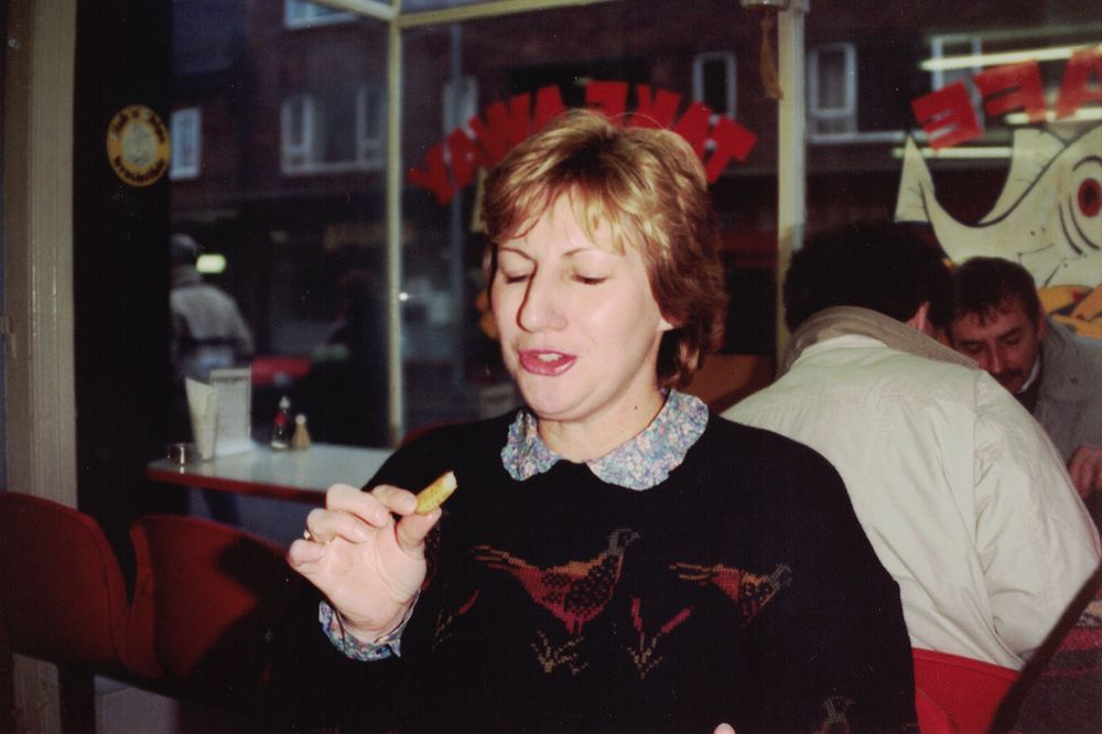 A woman with a dirty blonde bob and wearing a dark jumper with grouse (?) on it sits at a table in a fish and chip shop and holds a half-eaten chip.