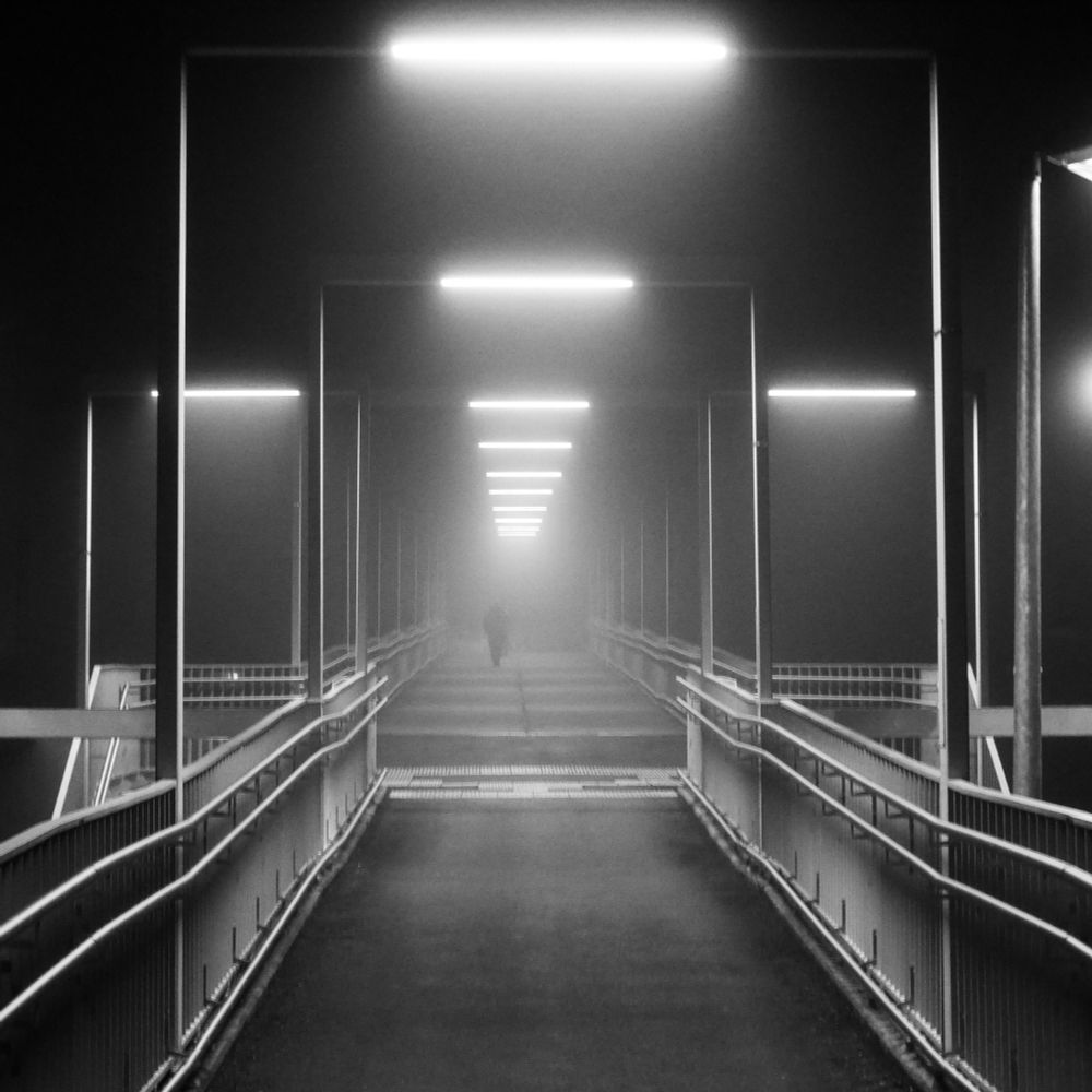 schwarz-weiß Foto einer zugangsrampe zu einem Bahnsteig von unten nach oben fotografiert. die Rampe ist sehr lang und mit portalartigen Lampen beleuchtet. das ganze ist in Nebel gehüllt und im fernen Hintergrund sieht man eine Person die Rampe nach oben laufen und im Nebel verschwinden 