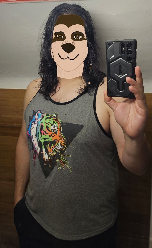 Mirror selfie of a person with a spectacled bear head cridely scribbled over the face. They have shoulder length wavy black hair and are wearing a gray tank top with a colorful tiger head design on it. The background is wood wainscoting and then white plaster above