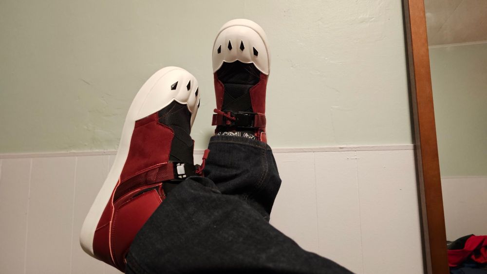 Picture of red shoes with wolf designs on the toe propped up against a pale green and white wall. The person wearing the shoes is also wearing dark blue jeans