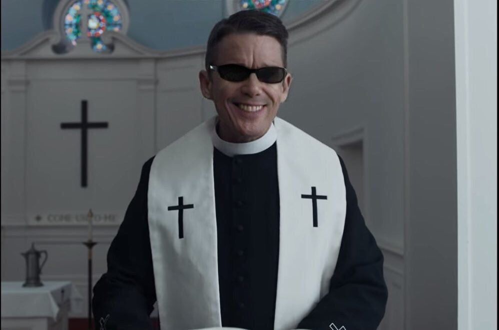 Reverend Toller rocking wraparound sunglasses and smiling effusively. 