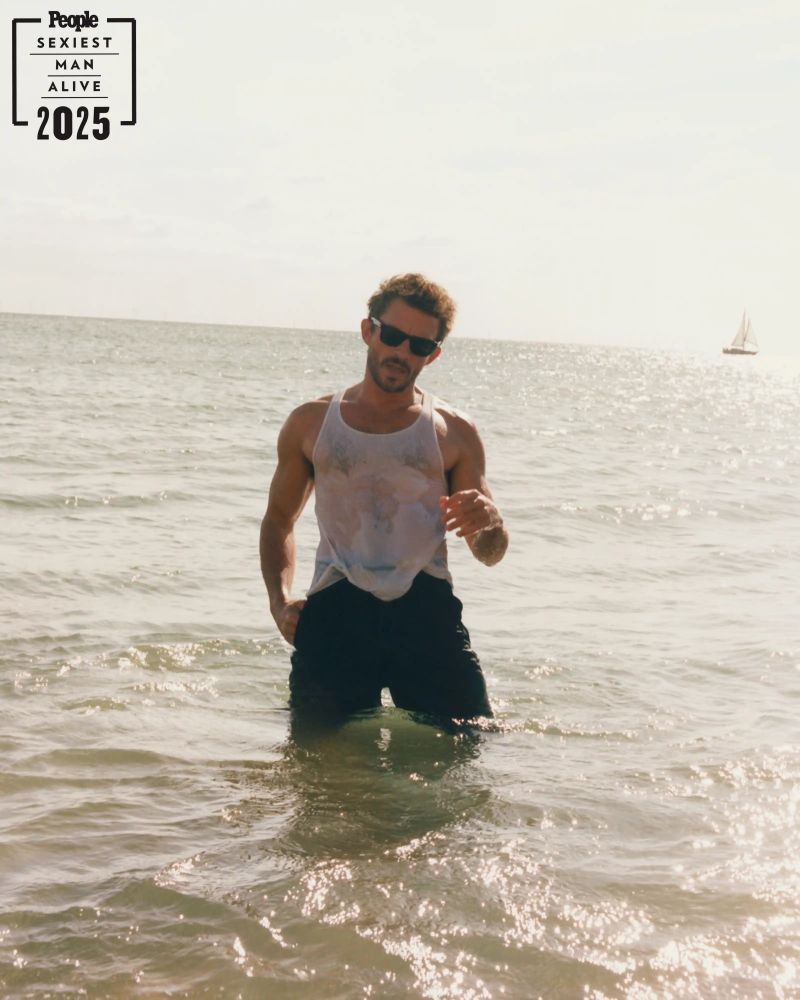 Foto de Jonathan Bailey para la edición de 'Sexiest Man Alive' de la revista People. Se ve a un hombre musculado, moreno y con barba que lleva puestas unas gafas de sol, una camiseta blanca de tirantes y unos pantalones negros. Está metido en el mar. El agua le llega a los muslos. La camiseta blanca está mojada y se le marcan los músculos.