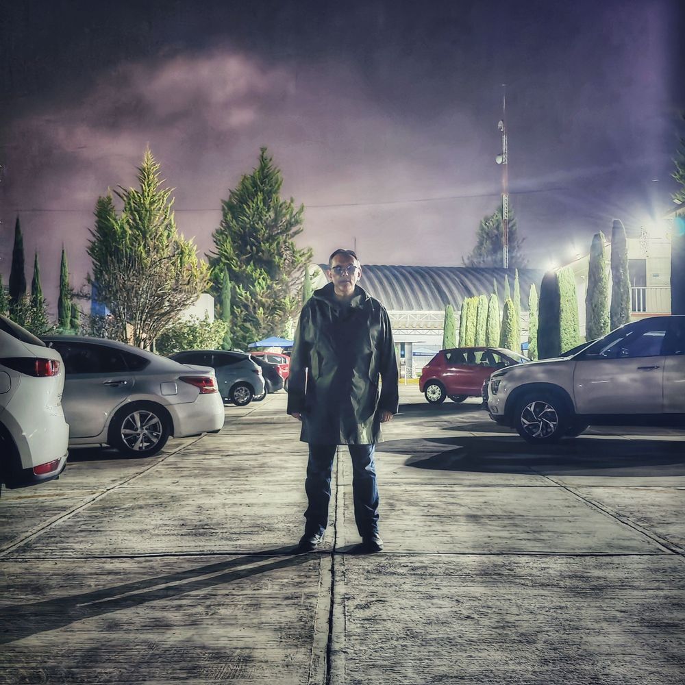 Un hombre vestido de gabardina verde está de pie en un estacionamiento. Autos de varios colores lo flanquean. El cielo nocturno es púrpura brillante.