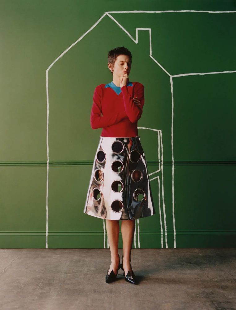 A white person with short brown hair (Emma Corrin) stands in front of a green wall that has a white outline drawing of a house on it. They are wearing black heels, a silver metallic knee length skirt with large grommet holes in it al over, and a red long sleeve polo with a blue collar.