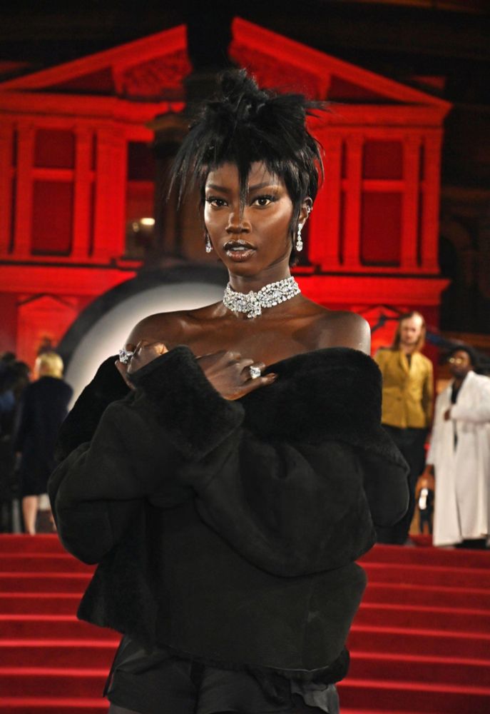 Anok Yai at The Fashion Awards 2024 in London on the red carpet stairs wearing a black fur shawl top that reveals bare shoulders to show off the diamond choker on her neck and a wild pointed jagged updo