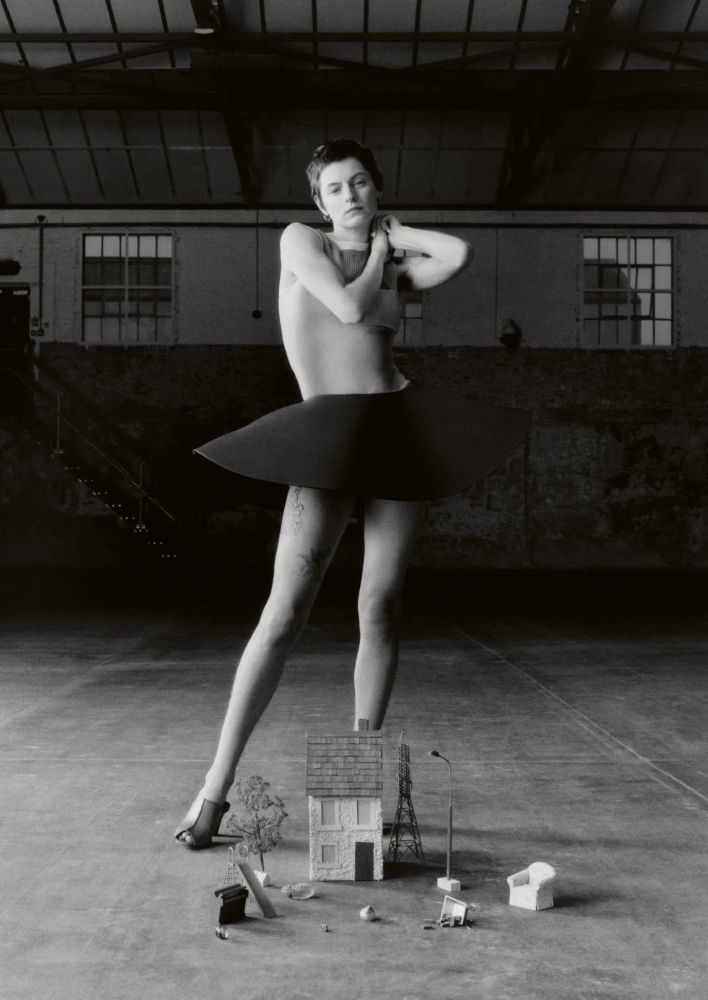 A white person with brown short hair (Emma Corrin) stands in the center of the frame in an empty studio behind a tiny set of miniature furniture on the lawn of a miniature house. They are wearing a flesh toned sheer sleeveless top that shows a white bralette with a very wide brim hat silhouetted skirt and cream mule shoes.