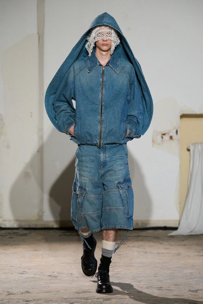 A runway model wearing a jean jacket that wraps up over onto the top of his head like a shelled hood over white lace and long jean shorts with cargo pockets and black leather boots
