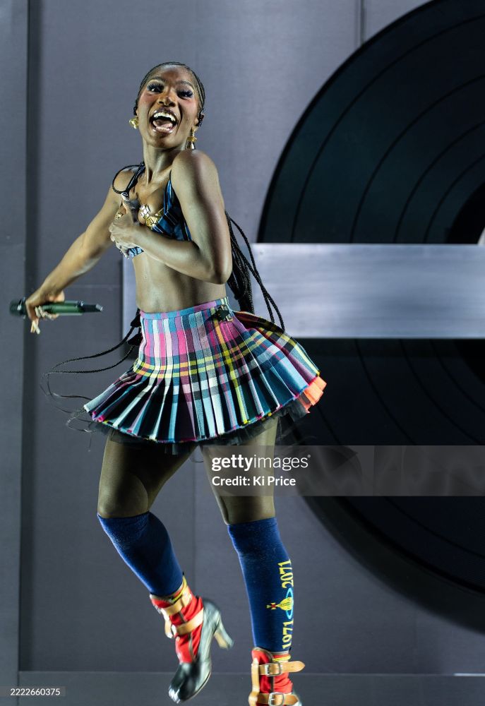 Doechii performing in full Vivienne Westwood (alongside her dancers) at glastonbury hands over her chest where she's wearing a navy blue bra, Westwood planet cross logo pasties, a rainbow colored plaid schoolgirl pleated skirt, knee high blue socks with the planet design on the outsides, red, yellow and black heeled boots.