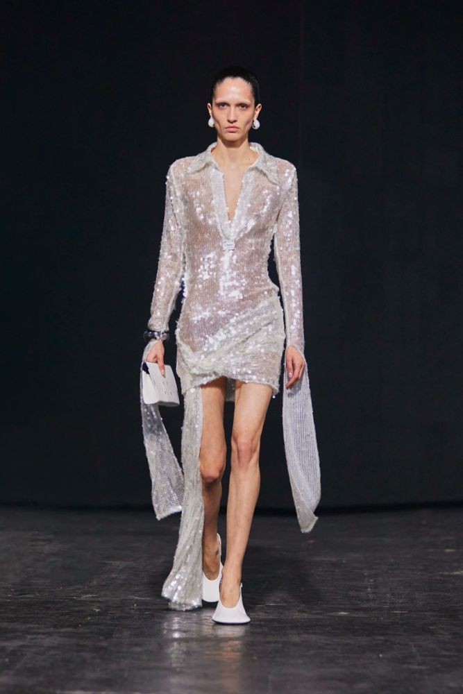 A runway model wearing a white sequined open collar dress with a draped asymmetrical fabric at the hips and long draping pieces from the wrists off the long sleeves.
