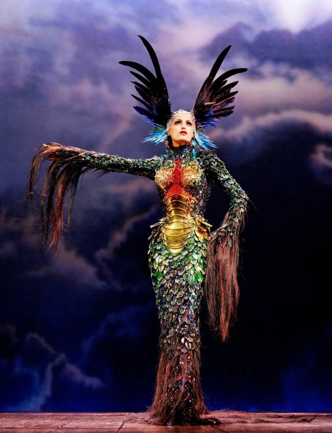 Yasmin Le Bon modeling a Thierry Mugler original scale and feather detailed gown with her arm out while the other has a hand on her hip. The headpiece is a large butterfly shape formed with feathers.