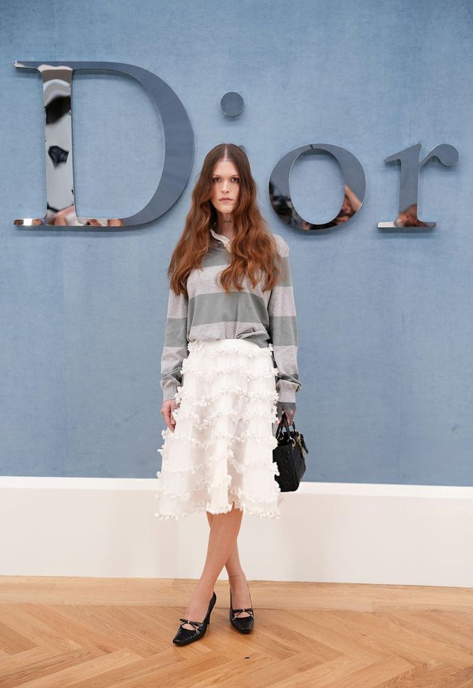 Ethel Cain wearing a grey and charcoal striped polo with a white tiered ruffled skirt and black heels with a black leather hand bag.