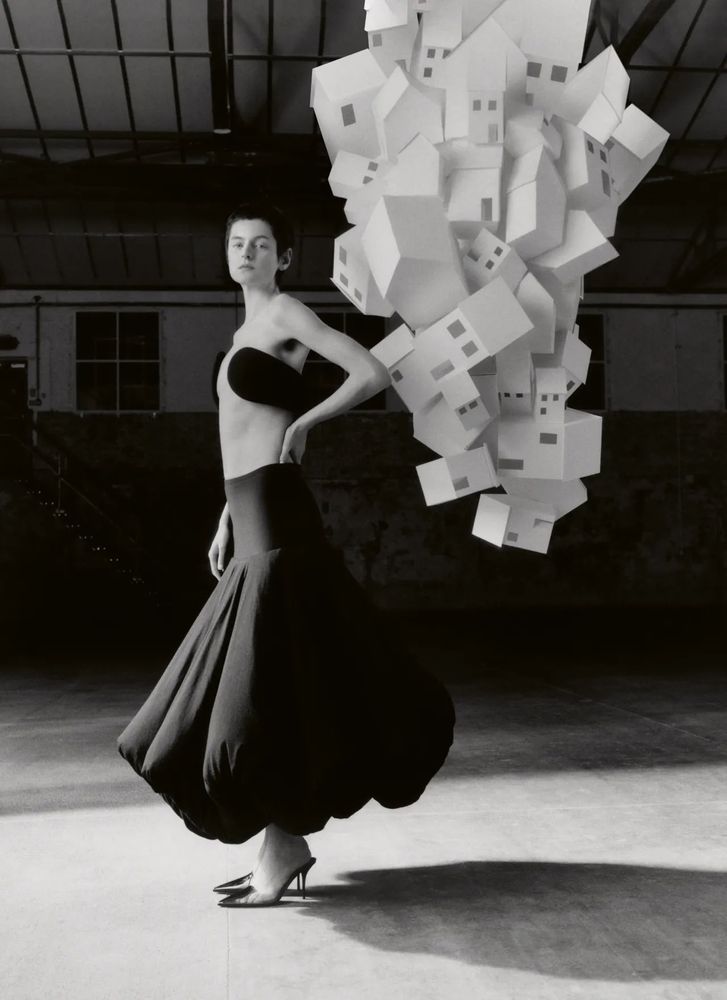 A black and white photo of a white person with brown short hair (Emma Corrin) posed to the side with a hand on their hip and a layered tornado behind them made up of paper white houses as they wear a black skirt that is ballooned out in sections at their ankle with two black strips covering their breasts meeting at the back and not the front middle with black heels in an empty studio