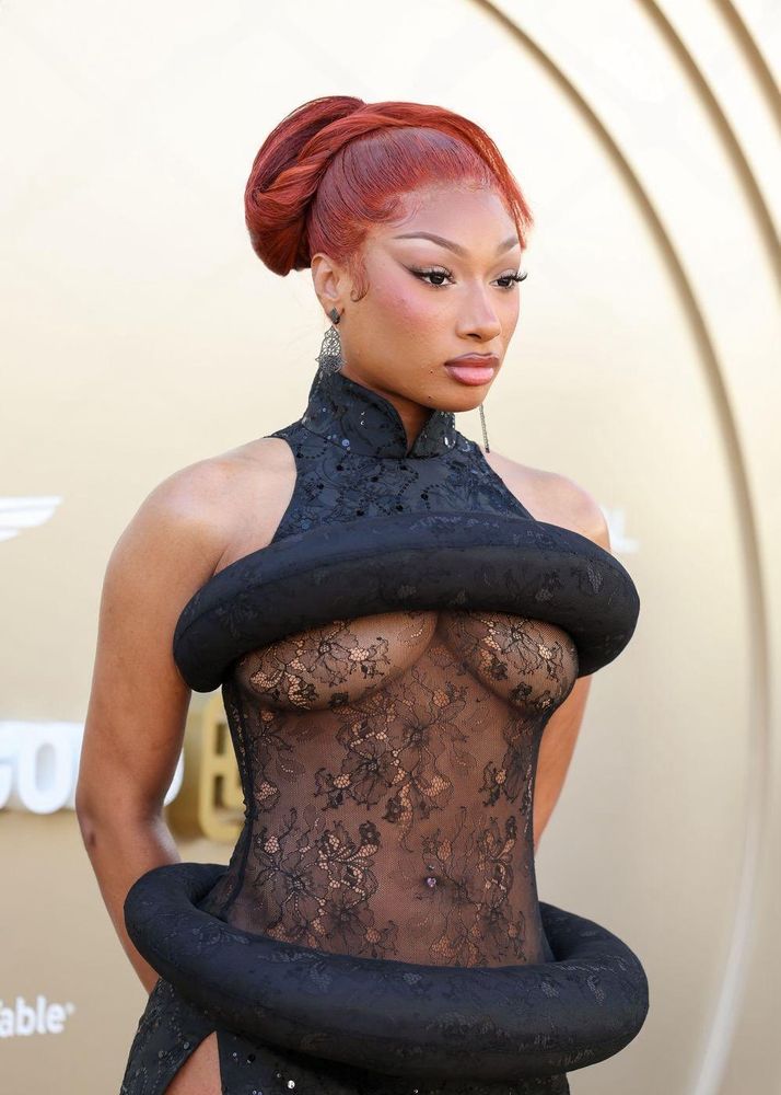 Megan Thee Stallion on a gold carpet for the Gold Gala wearing a Qipao style neckline that stops across her chest with the same fabric forming a circular hoop around her chest, her mid section is covered in lace which leads to another hoop with the Qipao style fabric and dress with slits on both sides revealing a peak of her strappy stiletto heels - all in black. She is posed with both hands on her hips with black stiletto shaped nails and red hair with a loose bun on the top of her head cascading off on either side.