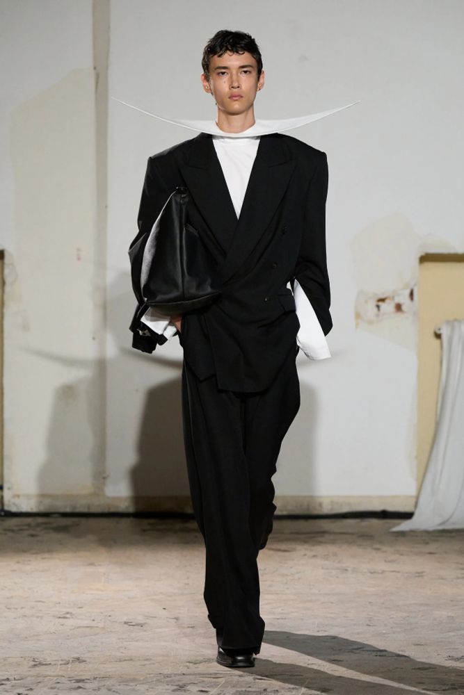 A runway model with a black suit and white shirt, the same style of sleeve cuffs as the first pic with an oversized u shaped collar the size of the shoulders.