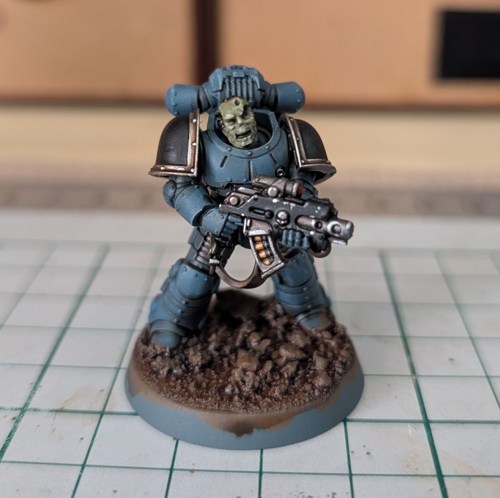 A Mark II space marine in light blue grey armour, with black shoulder pads and brass trim. The face is a horrible zombie green, with half decayed parts.