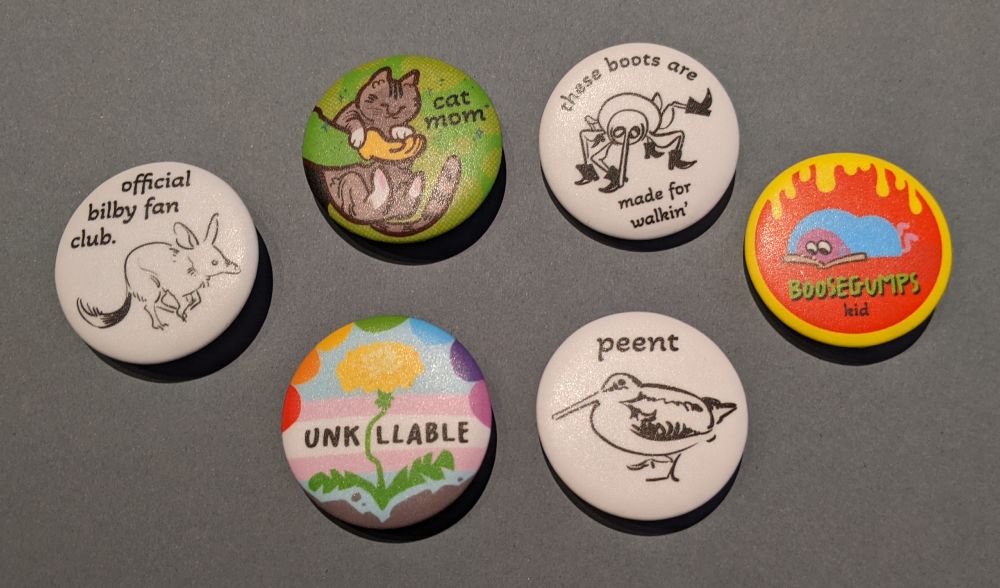 6 round button pins. Three are black and white and three are in colour. The black and white ones feature animals. "official bilby fan club." With a drawing of an australian bilby. "peent" with a drawing of an american woodcock. "these boots are made for walkin'" with a weevil wearing black cowboy boots. The colour ones are more varied. "cat mom" with a hand holding a grey cat with black stripes, the person is wearing green. "Boosegumps kid" with a pink cartoon person huddled under a blue blanket, reading a book. They are on a red background with dripping yellow design like the cover of Goosebumps books. "Unkillable" with a dandelion growing out of concrete, the background has rainbow petals and the trans flag colours.