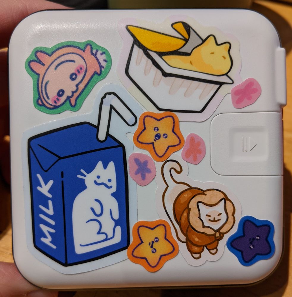 The other side of the battery, also covered in stickers. A blue juice box with milk and cat design, a custard cat peeking out of yogurt cup, a cat wearing a hooded jacket, stars making faces, and a bunny in a pink onesie.