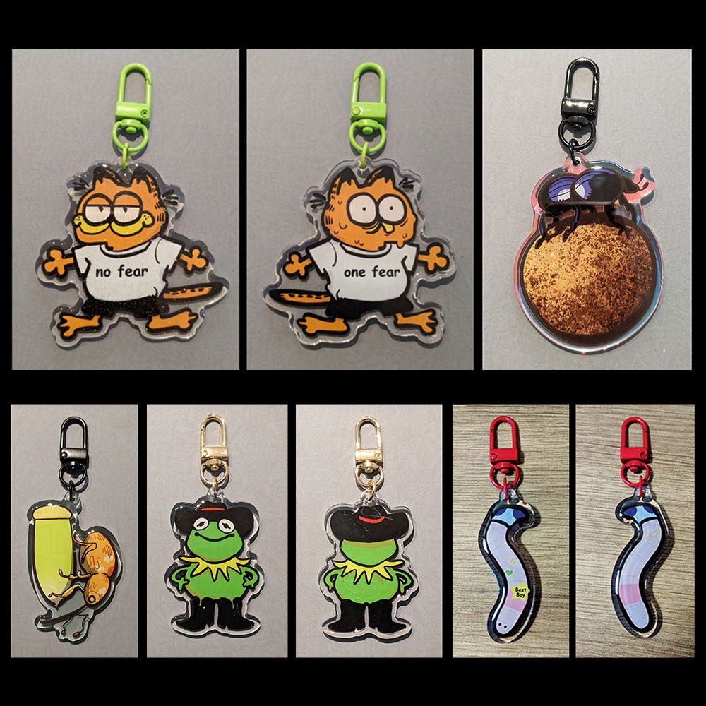 Acrylic keychain line up. Double sided Garfield keychain where he's wearing a white shirt and black shorts. Front side says "no fear" while he's smiling and relaxed, back side says "one fear" and he's wide eyed and sweating. Holographic blue and black dung beetle perched on a dung ball with black clasp. Acorn weevil perched on a green acorn with black clasp. Cowboy Kermit wearing black cowboy hat and boots. He is smiling. The clasp is gold coloured. Pink worm wearing a blue sock and a star badge that reads, "Best Boy"
