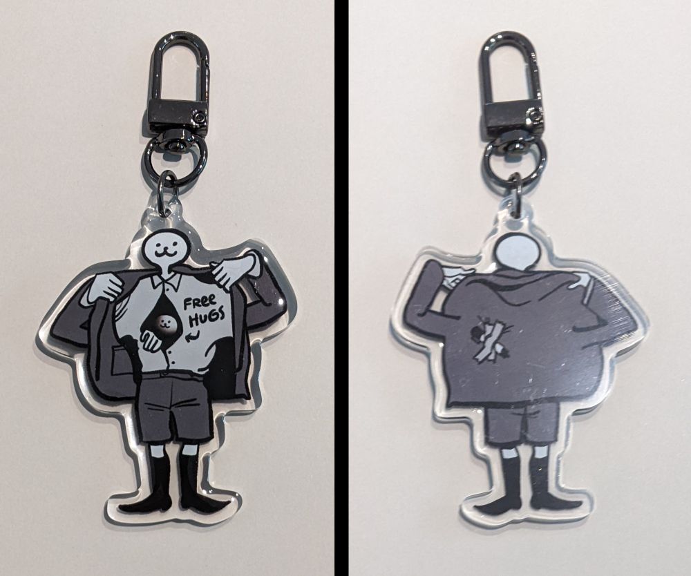2.5x2.5" acrylic keychain of a round headed figure wearing a grey suit. It holds open the jacket with long deformed arms to reveal a smiling face peeking out between the buttons of the shirt. On the shirt is written, "FREE HUGS" with an arrow pointing to the inside. A permanent marker is taped to the back of his jacket.
