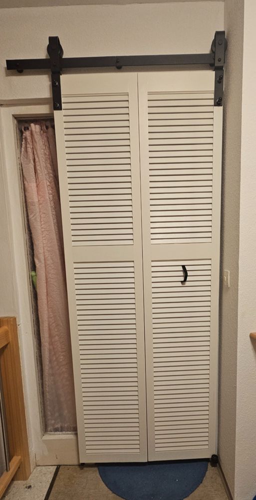 Weiße Lamellenklapptür mit schwarzen Band als Griff, von außen.